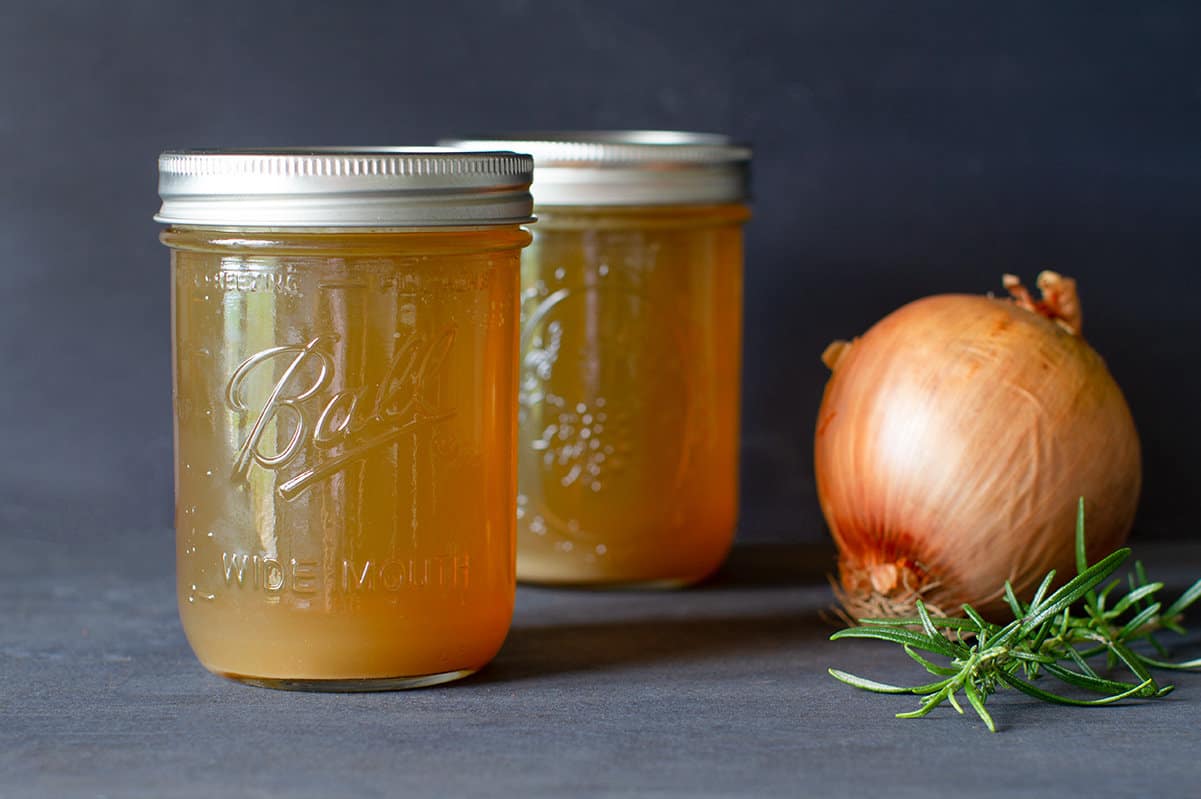 Two jars of golden lamb bone broth with an onion and sprig of rosemary.