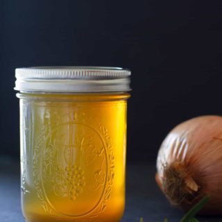 A jar of golden lamb stock with an onion and sprig of rosemary. Moody scene