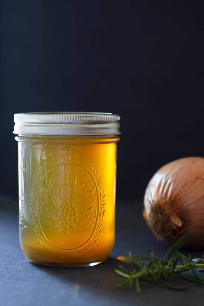 A jar of golden lamb stock with an onion and sprig of rosemary. Moody scene