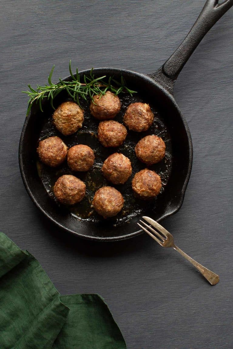 Greek lamb meatballs alongside a sprig of rosemary in a cast iron pan. A green tea towel is to the side.