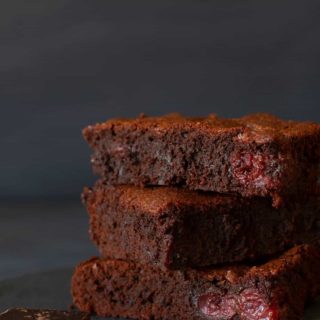 Three stacked squares of American brownies on a shingle next to a square of dark chocolate. Dark background.