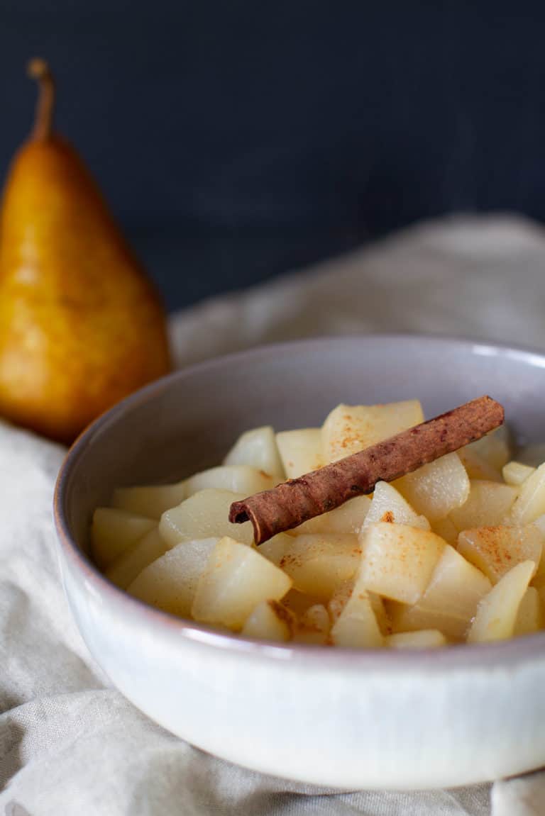 Close-up of a white bowl with stewed apples and pears with a cinnamon stick and a pear blurred in the background.