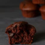 Close-up of a half triple chocolate muffin next to some chocolate drops and a stack of blurred muffins in the back.