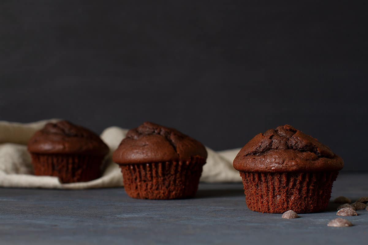 Three triple chocolate muffins in front of a linen tea towel. some chocolate drops are in the foreground.