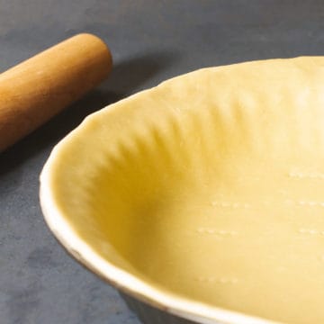 A 3 ingredient pie crust in a pie tin next to a rolling pin on a dark background.