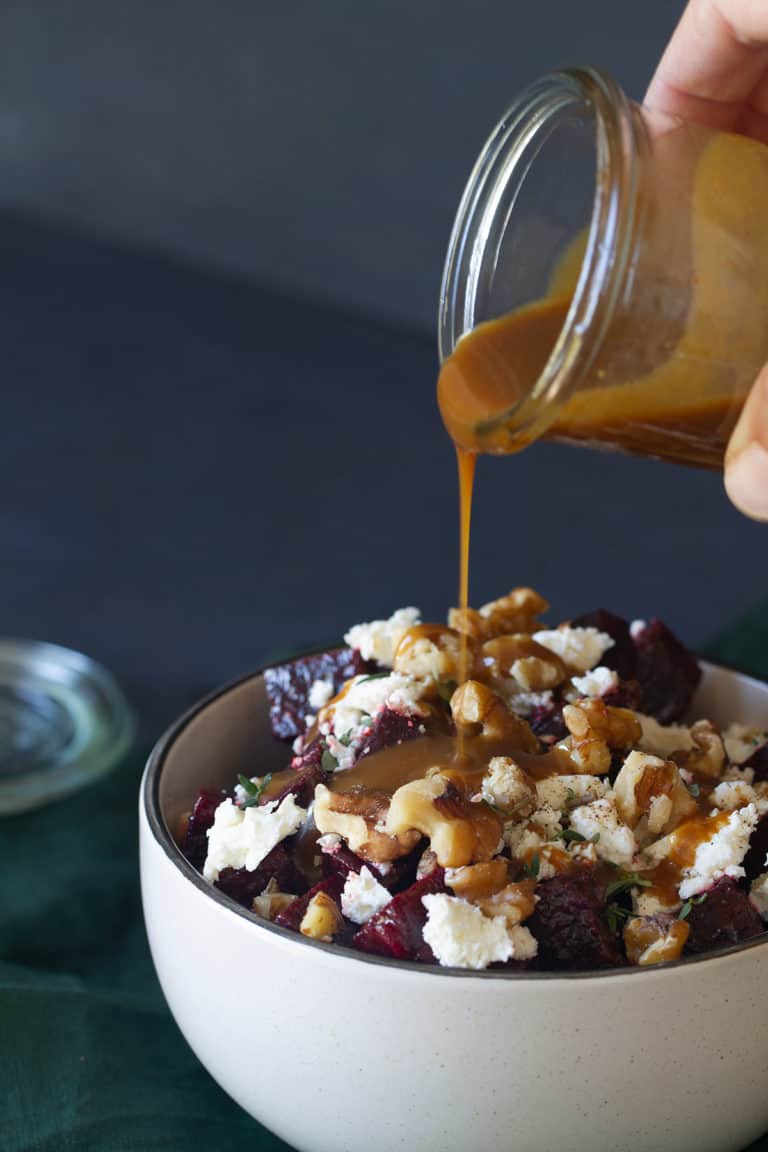 A bowl of roasted beetroot walnut and feta salad on a green tea towel. A jar of dressing is being poured onto the salad.
