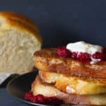 A stack of french toast on a black plate, topped with raspberries and yogurt. A brioche loaf is in the background.