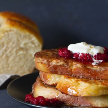 A stack of french toast on a black plate, topped with raspberries and yogurt. A brioche loaf is in the background.