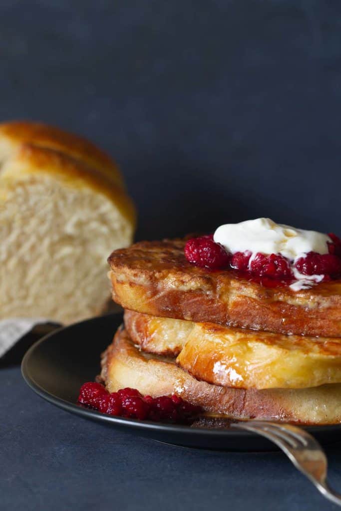 A stack of french toast on a black plate, topped with raspberries and yogurt. A brioche loaf is in the background.