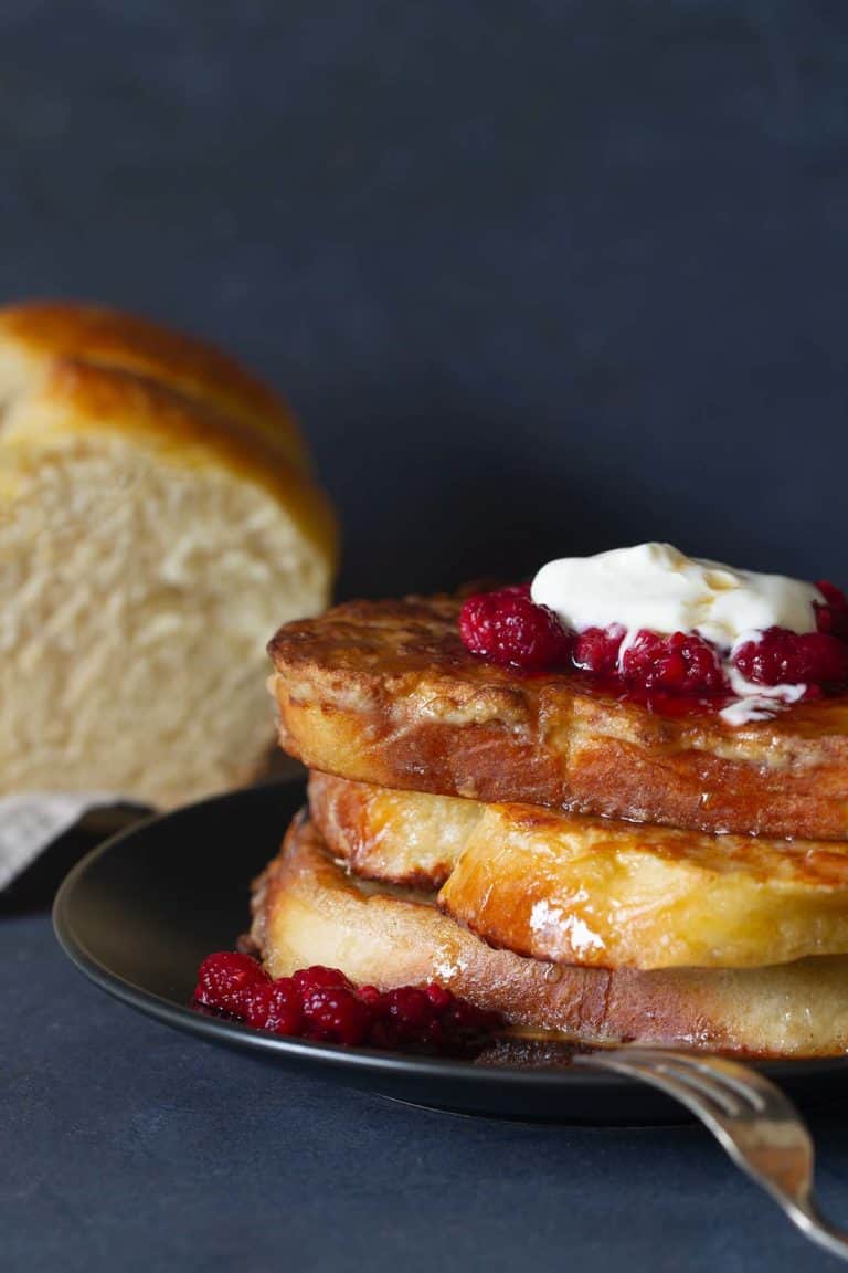 A stack of french toast on a black plate, topped with raspberries and yogurt. A brioche loaf is in the background.