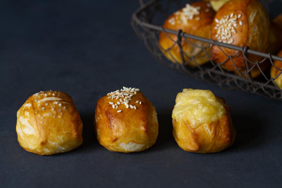 Three pretzel bites with different toppings in a row. A basket of more bites is in the background blurred out.