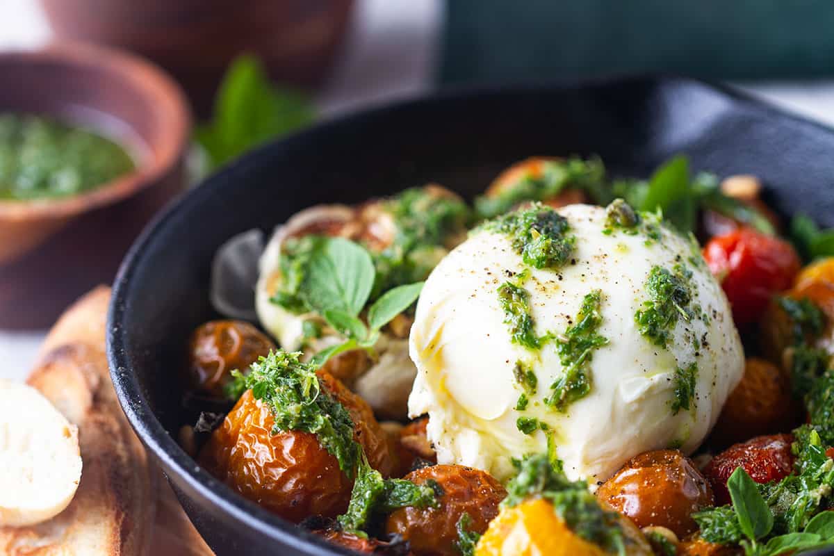 Close-up of a ball of burrata covered with salsa verde and next to slow-roasted tomatoes.