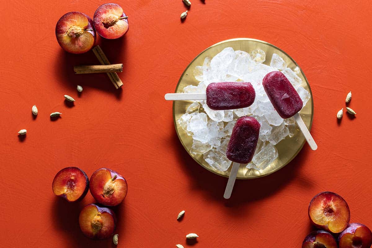 A golden tray with ice cubes and three gin plum ice blocks. Plums, and spices are next to it. Flatlay.