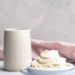 A bowl of whipped chantilly mascarpone next to a white ceramic mug, pink napkin and cinnamon sticks.