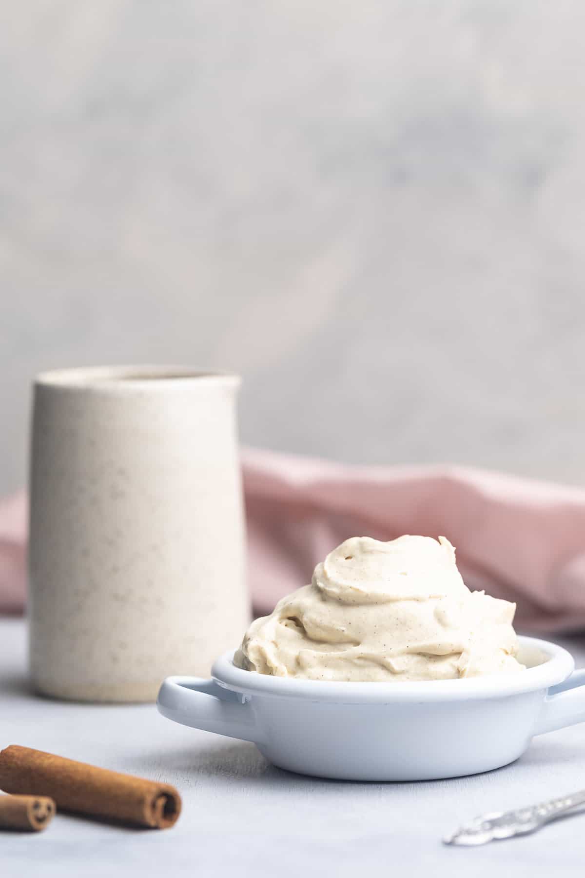 A bowl of whipped chantilly mascarpone next to a white ceramic mug, pink napkin and cinnamon sticks.