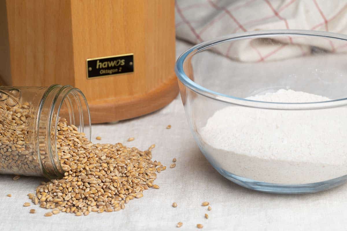 A jar of grains tipped over next to a bowl with flour and a grain mill in the background.