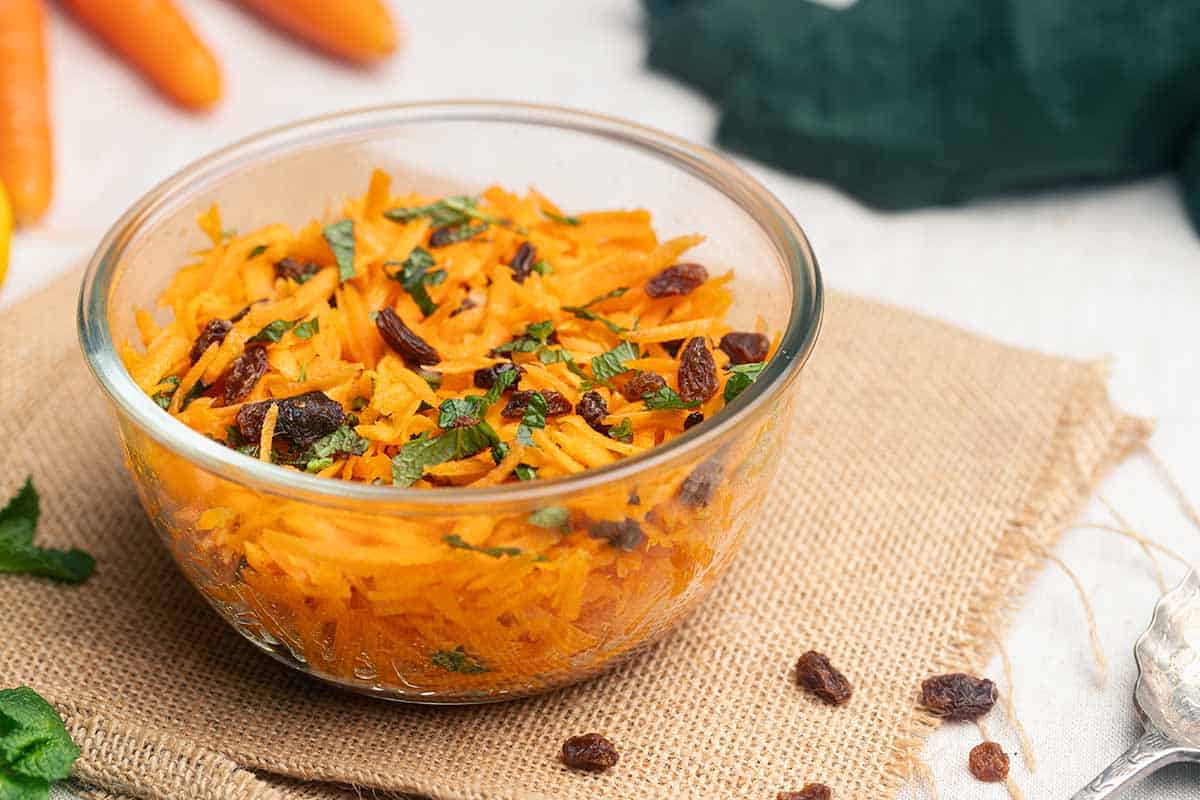 A glass bowl filled with raw carrot salad, garnished with mint and raisins.
