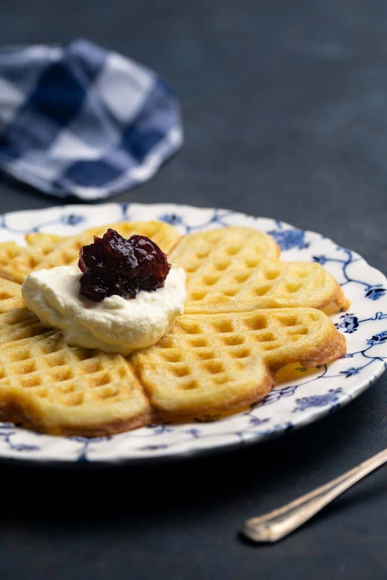 A thin and crispy Swedish waffle on a patterned plate, topped with cream and jam. Moody scene.