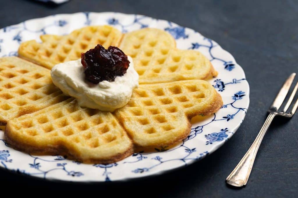 A thin and crispy Swedish waffle on a patterned plate, topped with cream and jam. Moody scene.