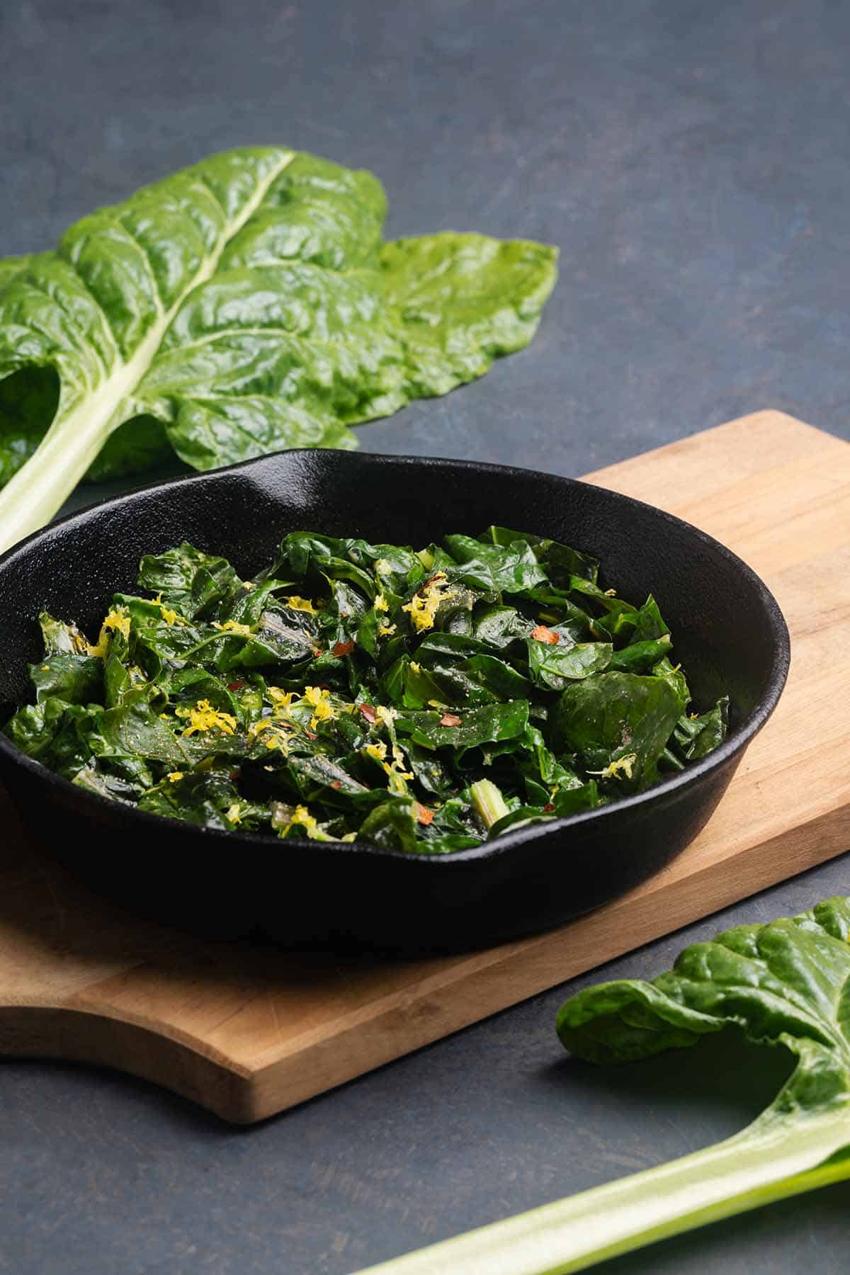 Sautéed silverbeet in a cast iron skillet on a wooden board next to silverbeet stems.