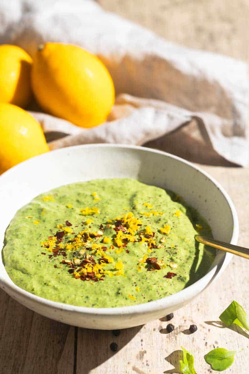 A bowl of creamy lemon pesto sauce next to lemons and a linen tea towel.