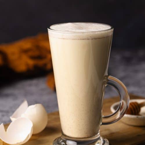 A tall glass cup filled with creamy egg milk on a wooden board next to egg shells.
