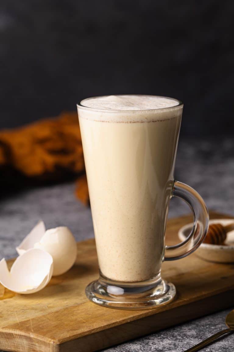 A tall glass cup filled with creamy egg milk on a wooden board next to egg shells.