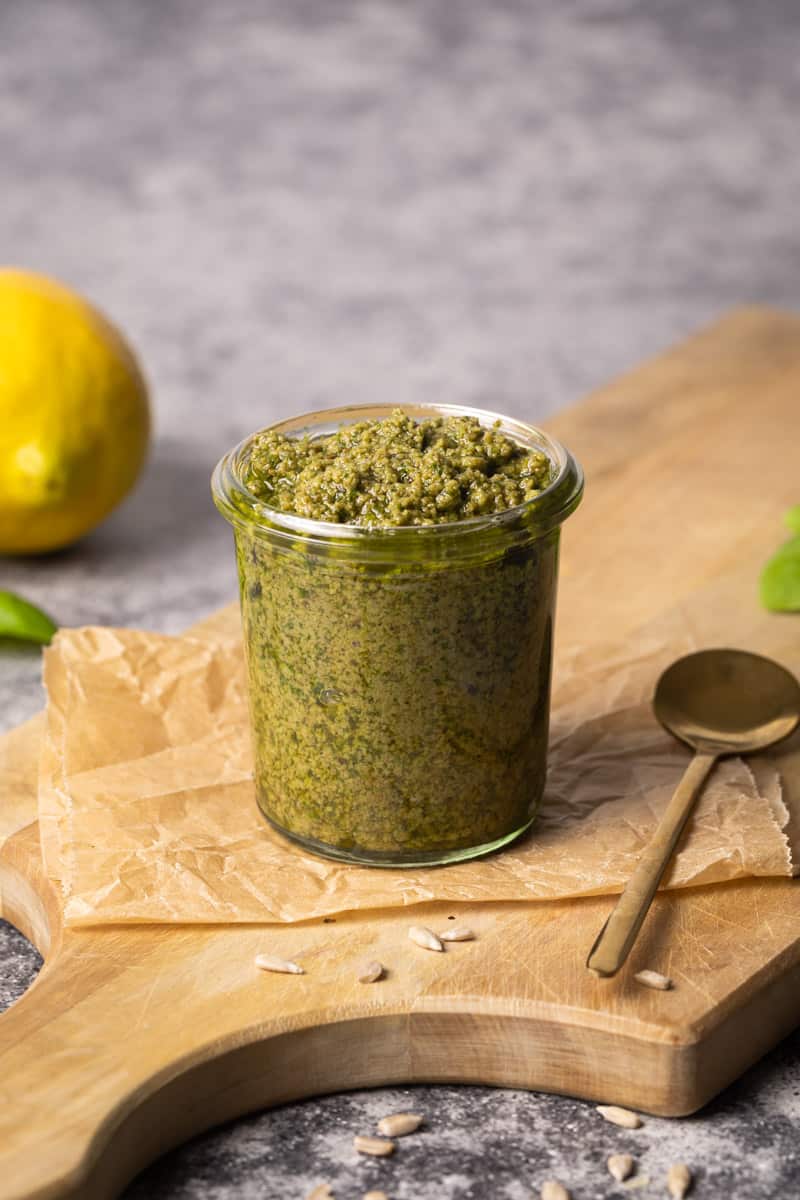 Basil sunflower seed pesto in small glass jar on wooden board next to sunflower seeds, a golden spoon, basil, and a lemon.