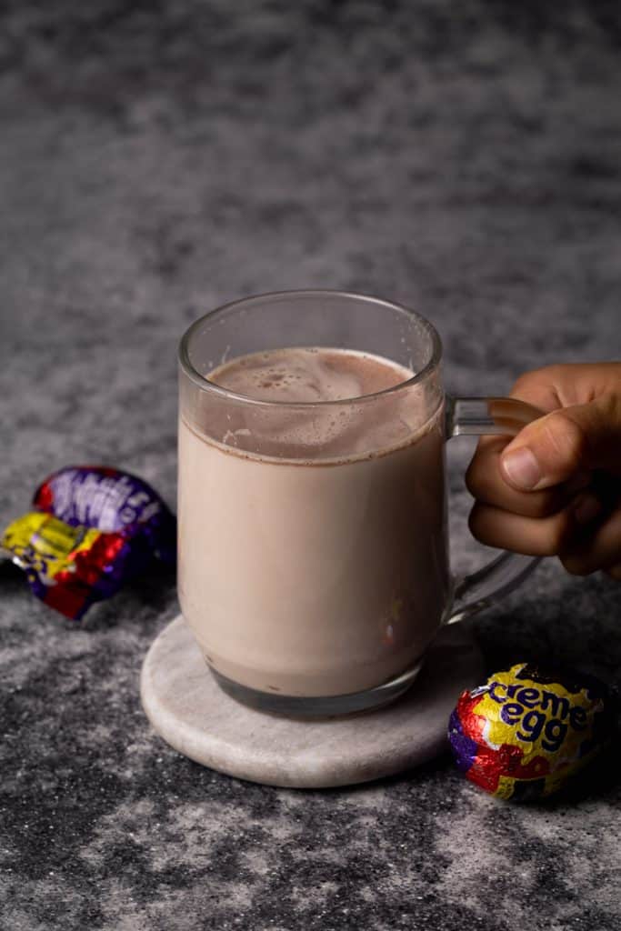 Cadbury creme egg hot chocolate in a glass cup next to creme eggs.