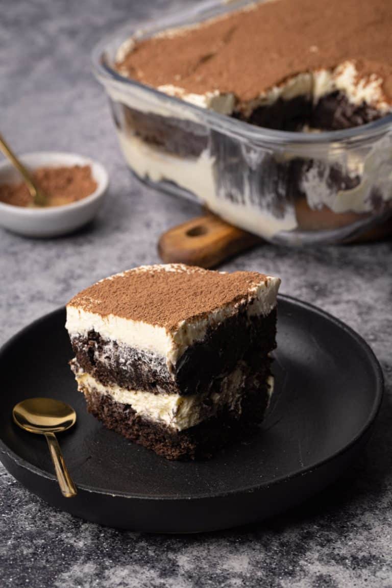 A slice of brownie tiramisu on a black plate next to the whole dish.