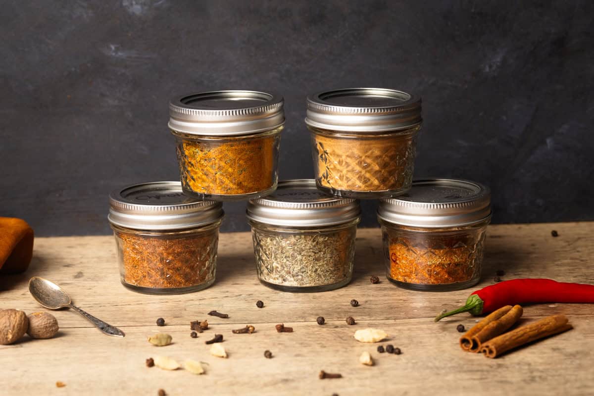 Five spice jars stacked up on a wooden background next to loose spices.