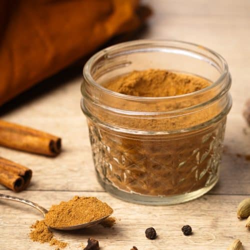 A jar of homemade Masala chai spice mix next to whole ingredients and a small spoon.