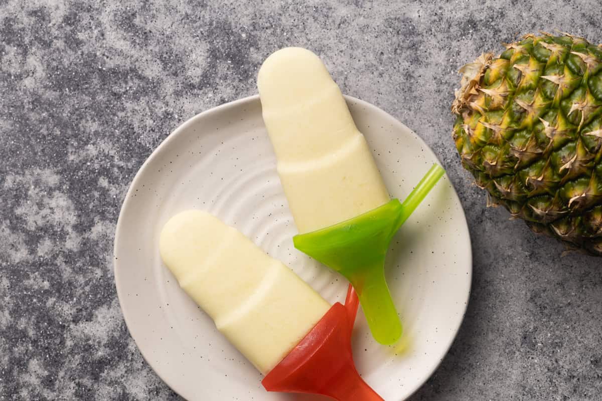 Two tropical popsicles on a white plate next to a pineapple.