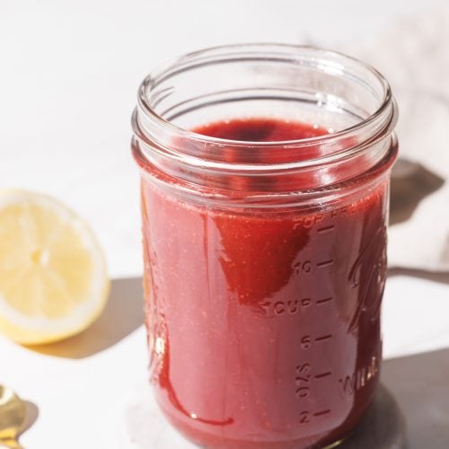 A mason jar filled with strawberry puree on a marble coaster next to a golden spoon and half a lemon.