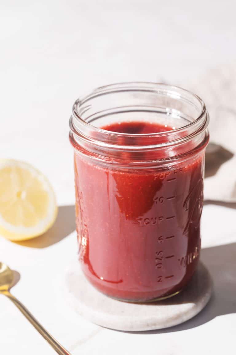A mason jar filled with strawberry puree on a marble coaster next to a golden spoon and half a lemon.
