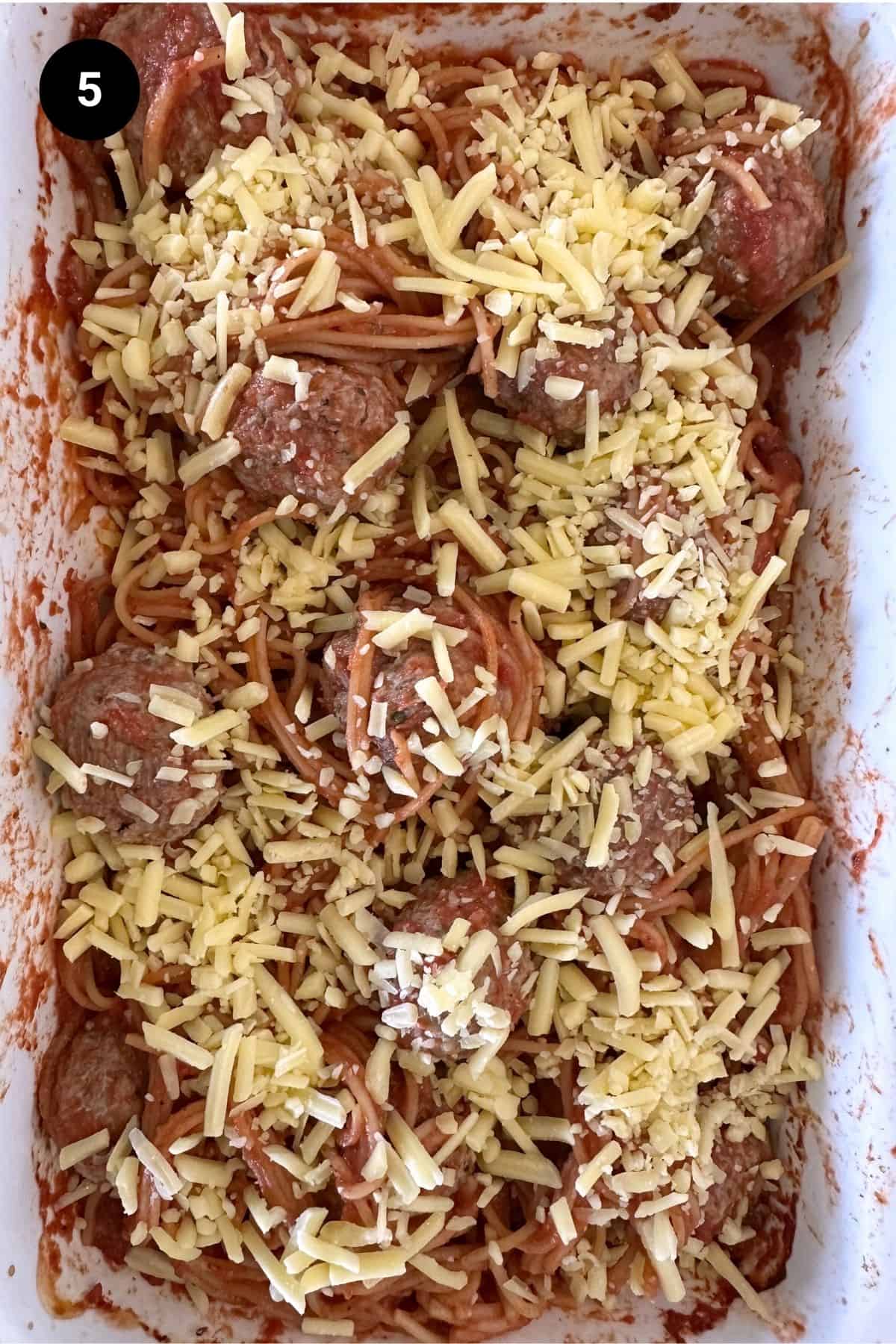 Spaghetti and meatballs before being baked in a white casserole dish, covered with shredded cheese.