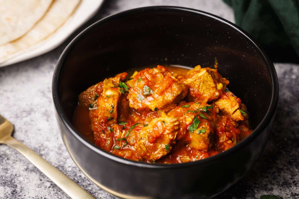 A black bowl filled with lamb curry next to naan bread and a green napkin.