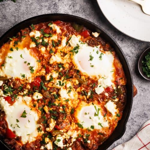 Sausage shakshuka in a cast iron pan next to a tea towel, plates, and parsley.