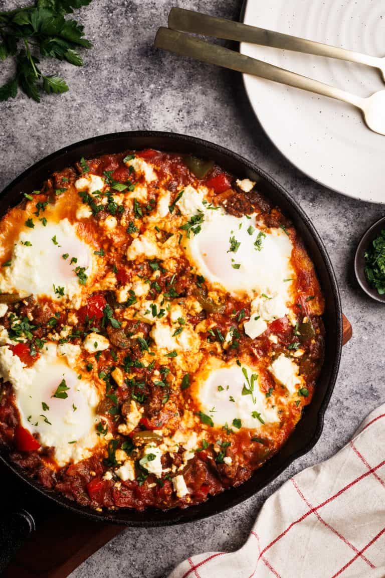 Sausage shakshuka in a cast iron pan next to a tea towel, plates, and parsley.