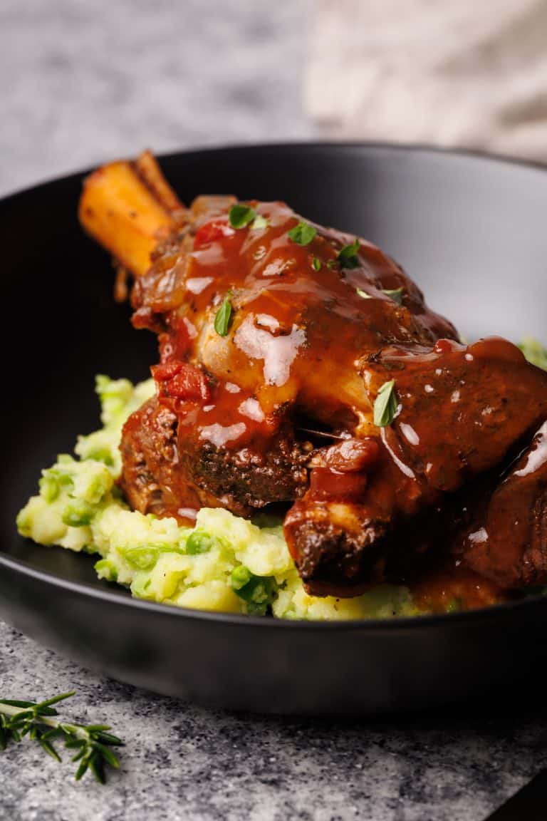 Lamb shanks on mashed peas and potatoes in a black bowl.