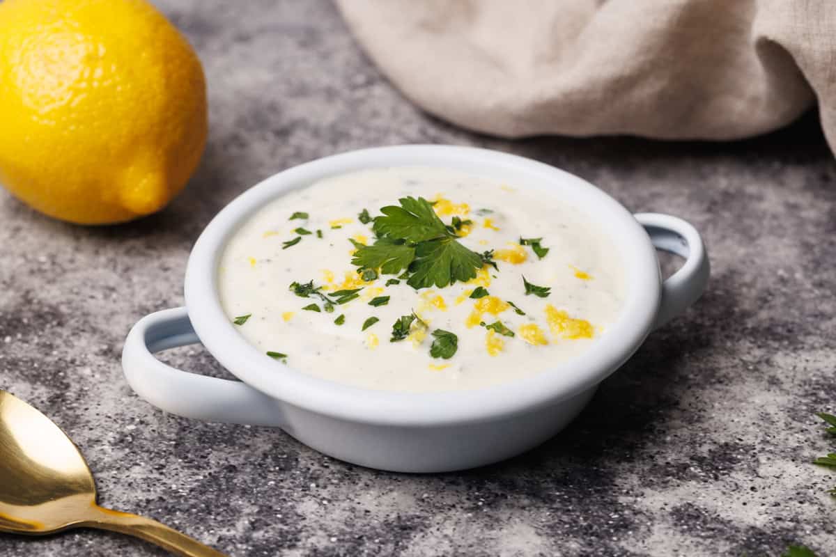 Lemon yogurt sauce in a small white enamel dish next to a lemon and natural linen tea towel.
