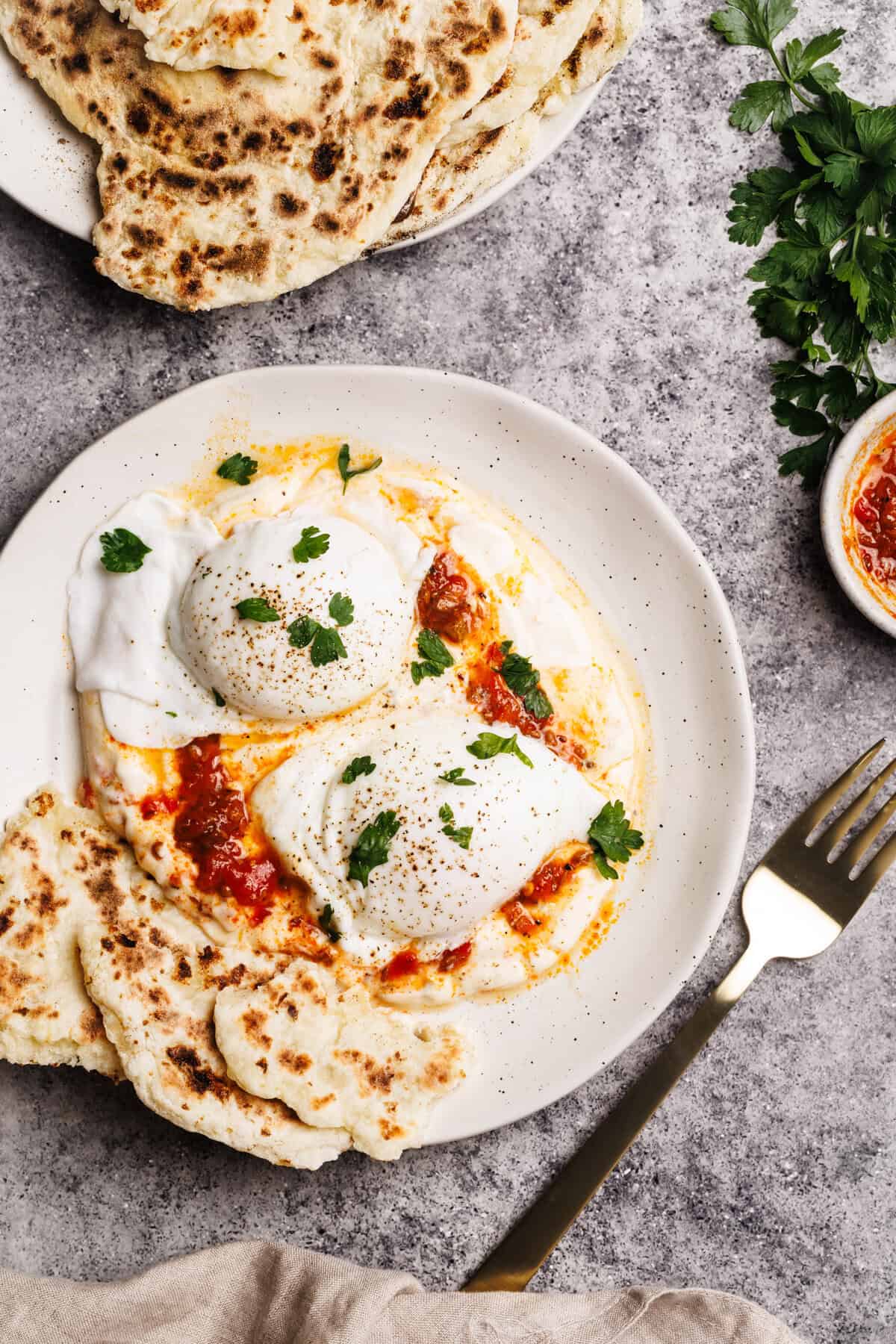 Turkish eggs with harissa on a white plate next to flatbread.