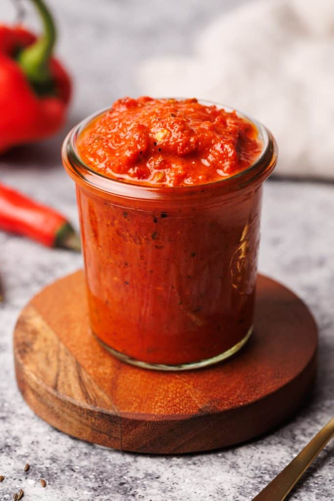A glass jar filled with harissa paste on a wooden coaster next to chilis.