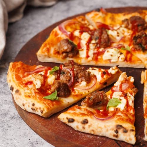 Cut lamb sausage pizza on a round wooden board.