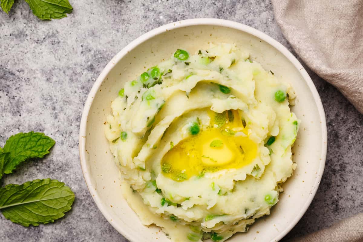 A cream bowl filled with minty mash on a grey background next to fresh mint leaves.