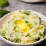 A cream bowl filled with minty mash on a grey background next to fresh mint leaves.
