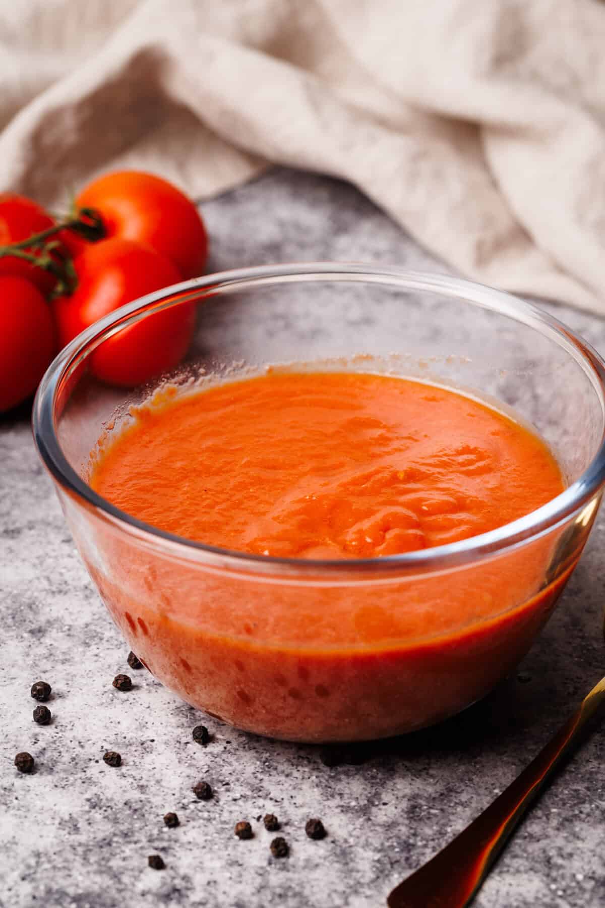 Pizza sauce in a glass bowl next to fresh tomatoes, a napkin, and black pepper.