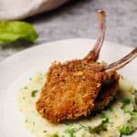 Two crumbed lamb chops in some minted pea and potato mash on a white plate.