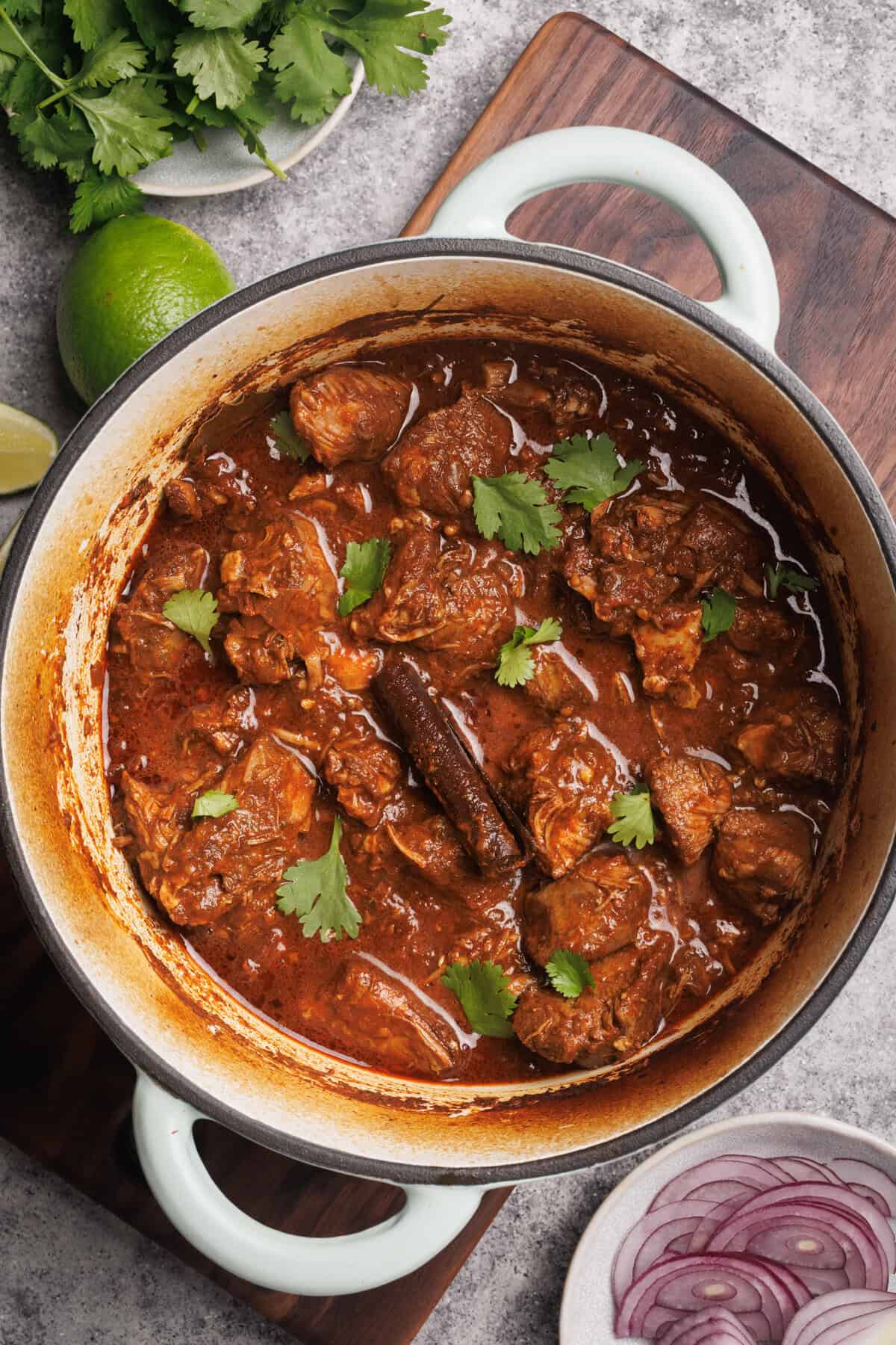 Lamb birria in a Dutch oven next lime, red onion, and coriander.