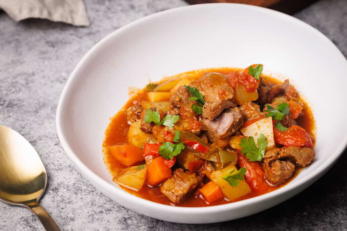 Hungarian lamb goulash in a white bowl next to parsley and a golden spoon.